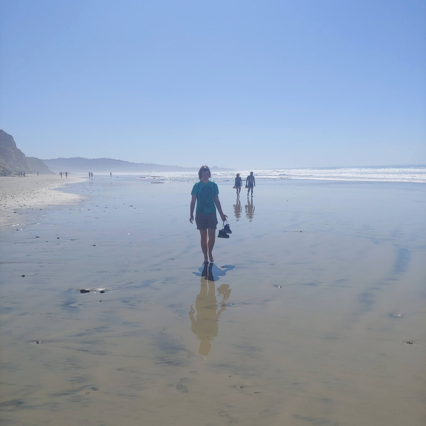 Carolyn walks on a beach carrying her hiking boots.